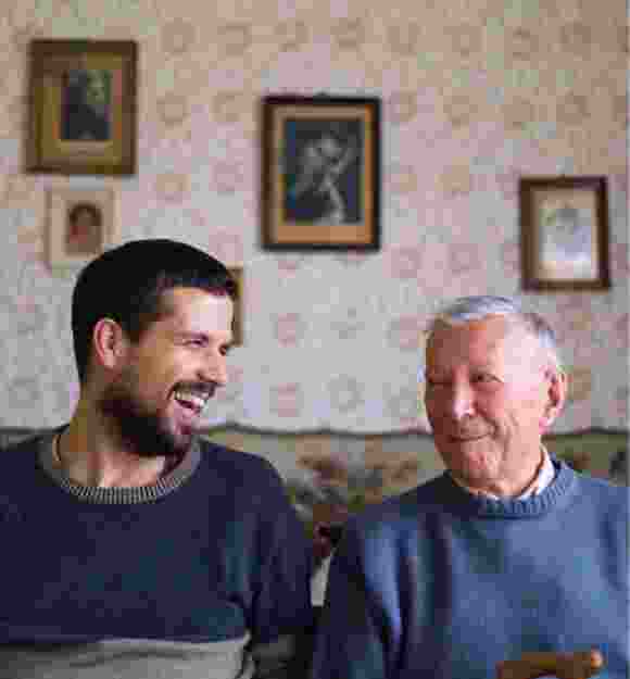 Two people, one adult & one older person, sitting on a couch looking at each other laughing.