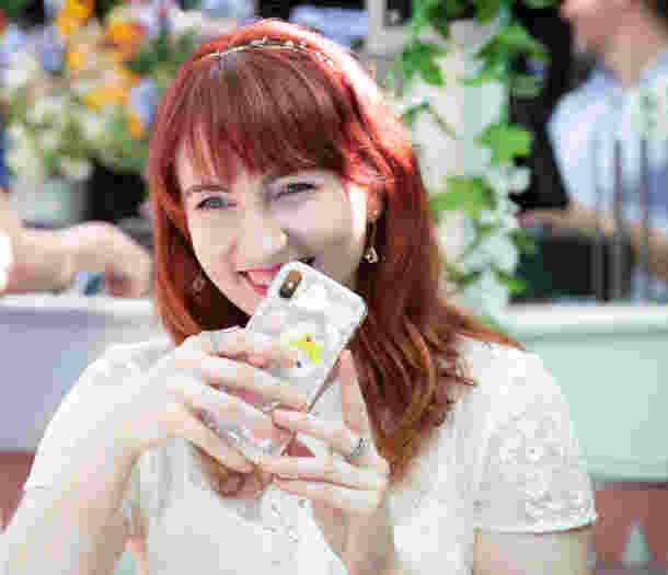 An adult who is outside at a cafe laughing. They have their phone up to their mouth and it is covering part of their mouth.