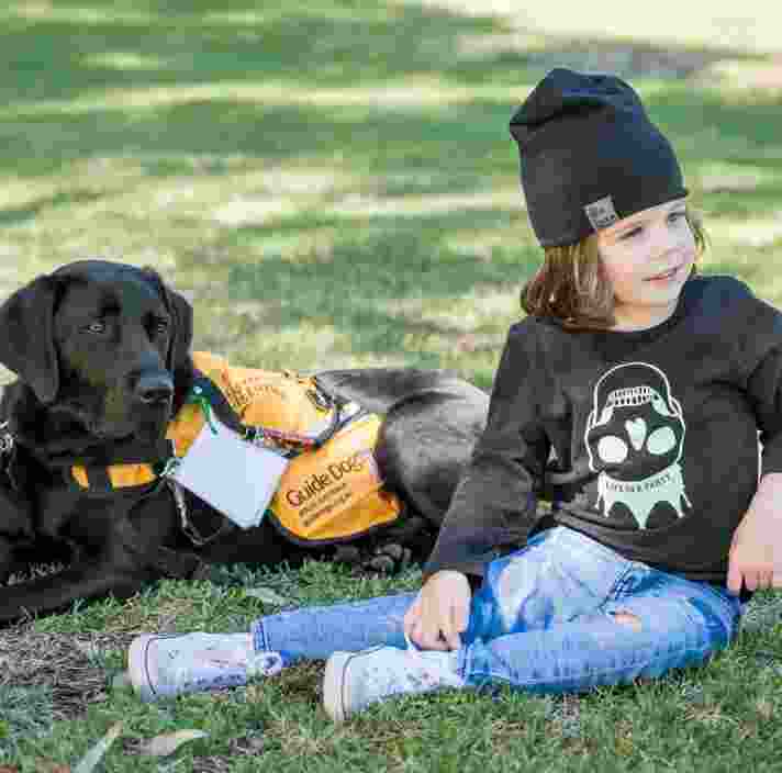 A young child sitting outside on some grass next to an Autism Assistance Dog. The dog is a black labrador and is wearing an Autism Assistance Dog jacket.