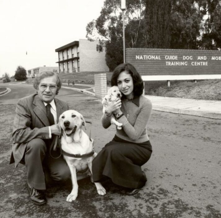 A 1940's black and white image of two people outside a Guide Dog Training Centre. One of the people in kneeling down next to a yellow labrador Guide Dog in harness and the other is holding a yellow ten week old labrador puppy. Both dogs and people are looking at the camera.