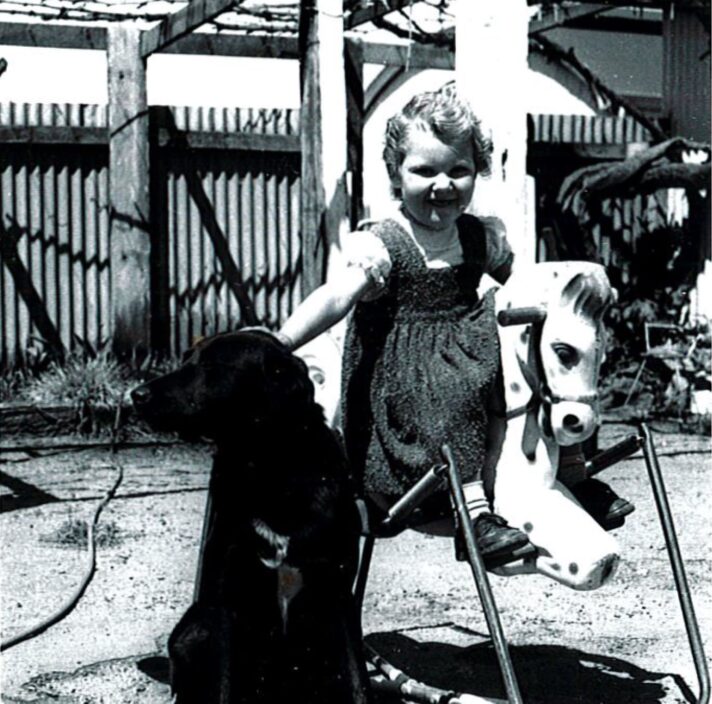 1940's Black and white photograph of a child riding a toy horse whilst patting a black labrador dog that is next next to them. The child has a smile on their face.