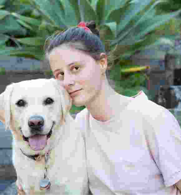 A person looking straight at the camera with their arm around a yellow labrador dog. They are seated at the bottom of a set of stairs.