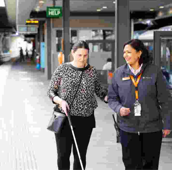 A person using their white cane at the train station while being guided by a train station attendant.