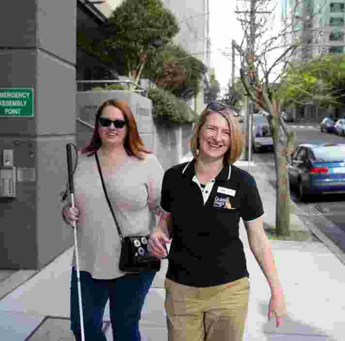 A person with their white cane walking down the street with a Guide Dogs staff member. Both people are smiling at the camera and the person with the cane is holding the staff member by the elbow.