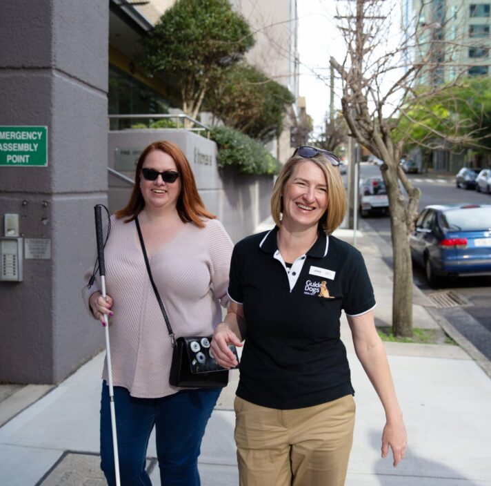A person with their white cane walking down the street with a Guide Dogs staff member. Both people are smiling at the camera and the person with the cane is holding the staff member by the elbow.