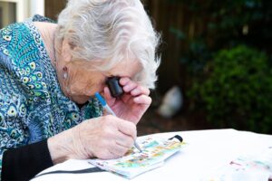 An older adult using a magnifier whilst painting, The person is painting onto a canvas.