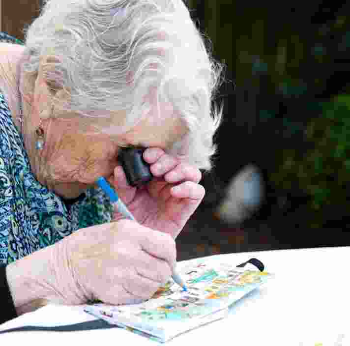 An older adult using a magnifier whilst painting, The person is painting onto a canvas.