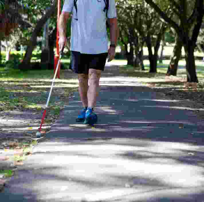 A person using their white cane while walking down a path in a park.