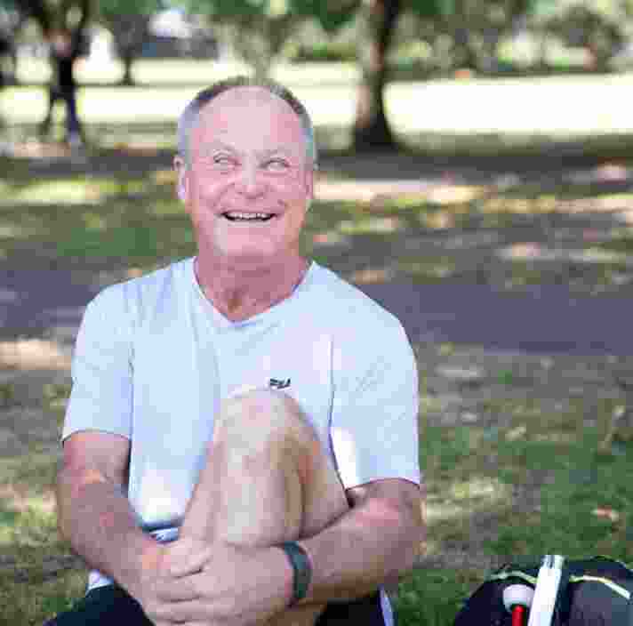 A person sitting outside on the grass with a smile on their face looking at the camera. They have one leg bent with their arm wrapped around their knee and a white cane next to them.