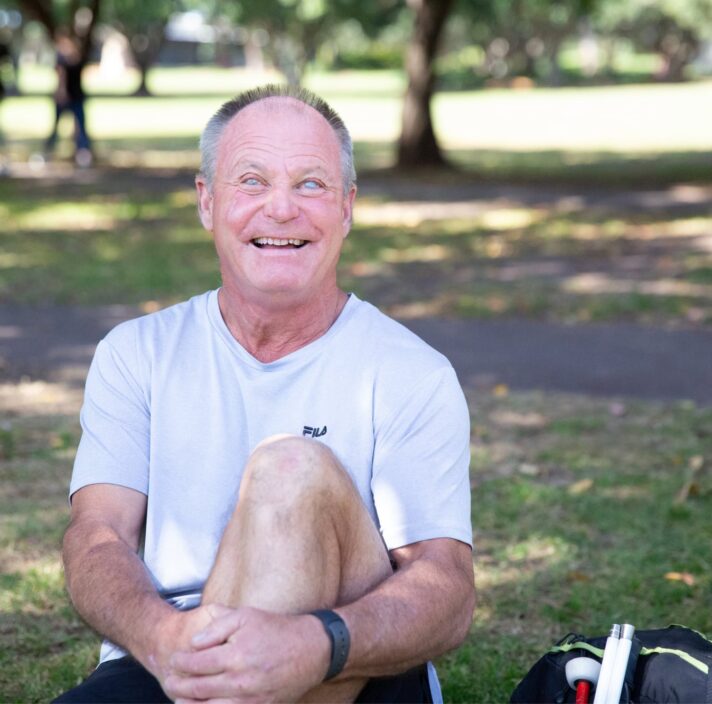 A person sitting outside on the grass with a smile on their face looking at the camera. They have one leg bent with their arm wrapped around their knee and a white cane next to them.