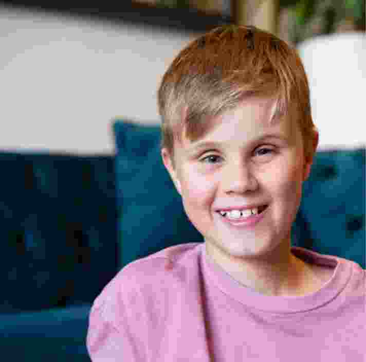 Close up of a young child with a big smile on their face. They are seated on the ground with a couch in the background.
