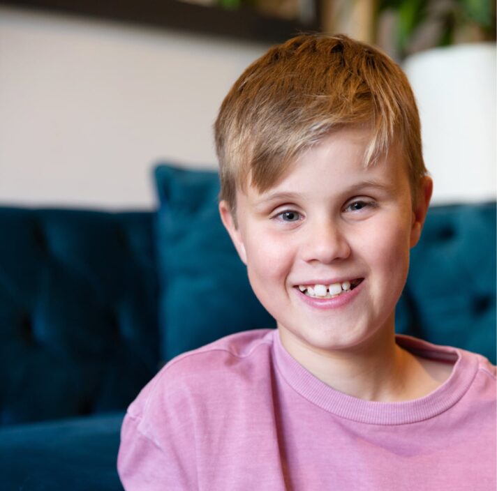 Close up of a young child with a big smile on their face. They are seated on the ground with a couch in the background.