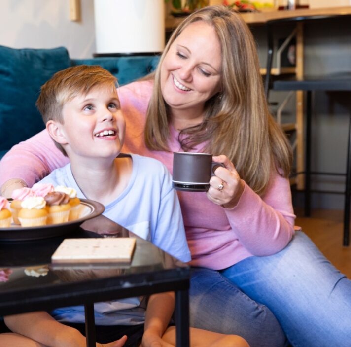 Mother sitting on the ground next to her child and they are both smiling. The mother is holding a mug and there is a plate of muffins on the table in front of them.