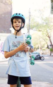 Young child wearing a helmet, holding a skateboard and standing outside in the city streets. The child is looking at the camera and smiling.