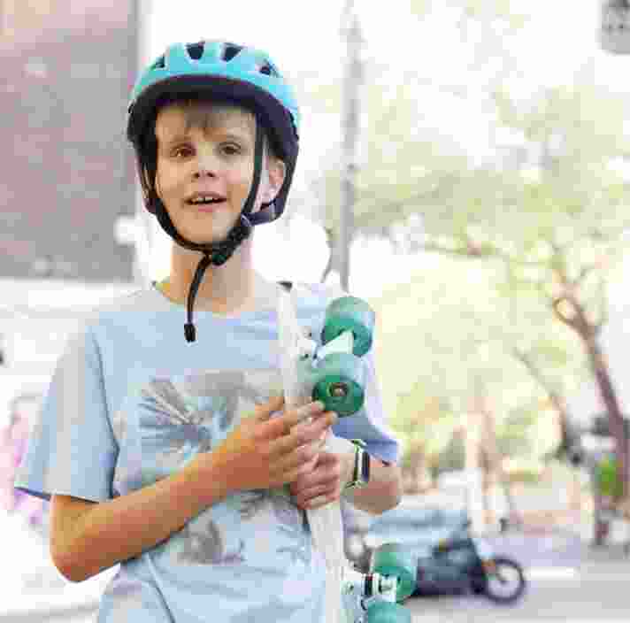 Young child wearing a helmet, holding a skateboard and standing outside in the city streets. The child is looking at the camera and smiling.