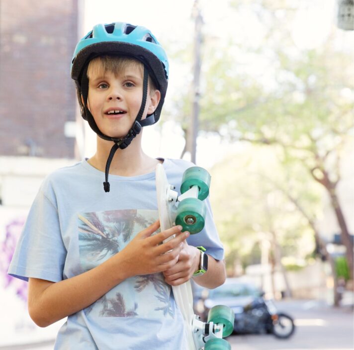 Young child wearing a helmet, holding a skateboard and standing outside in the city streets. The child is looking at the camera and smiling.