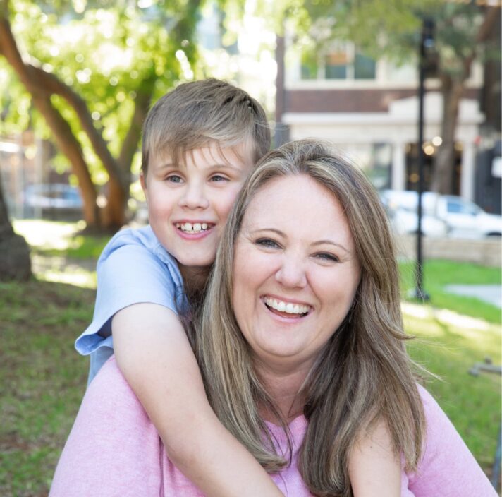 Young child with their arms around their mothers neck and they are both looking at the camera smiling. They are seated on grass outside.