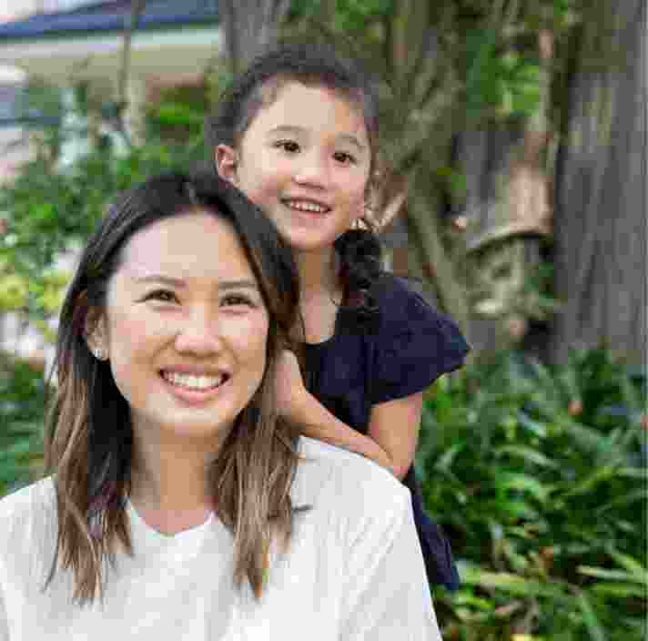 A mother and young child outside smiling at the camera. The young child is standing behind the mother with their hands on her back.