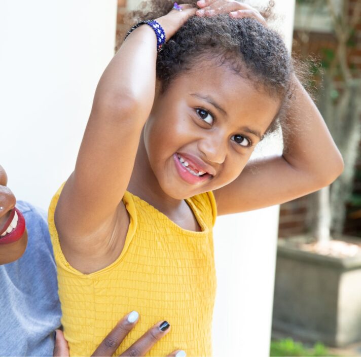 A young child smiling at the camera while their mother looks at them from behind. The mother is smiling at their child.