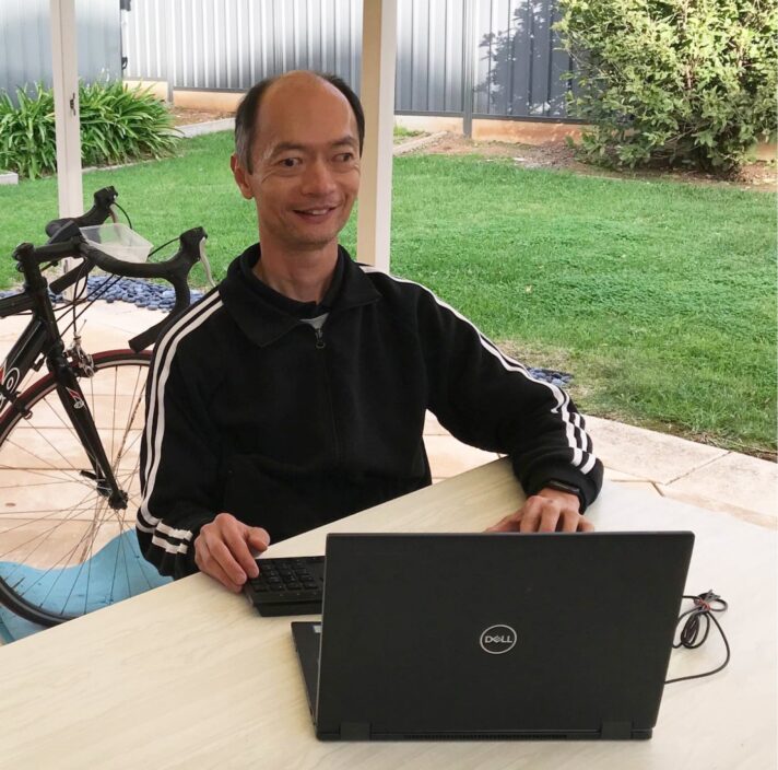 A person sitting outside at a table with their laptop. The person is smiling at the camera.