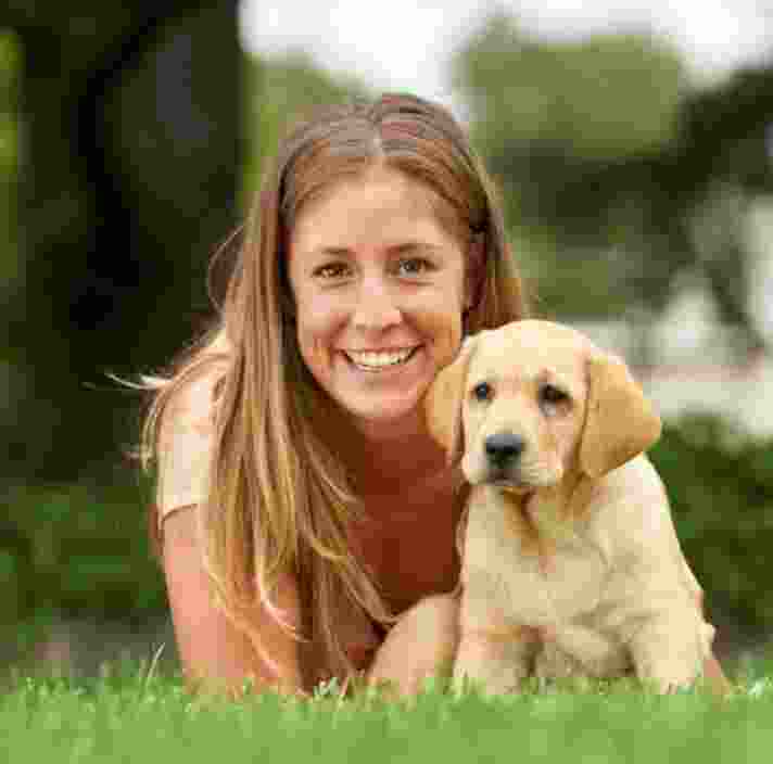 A person laying on some grass with an eight week old yellow labrador puppy. The person is smiling and looking at the camera. The puppy is also looking at the camera.