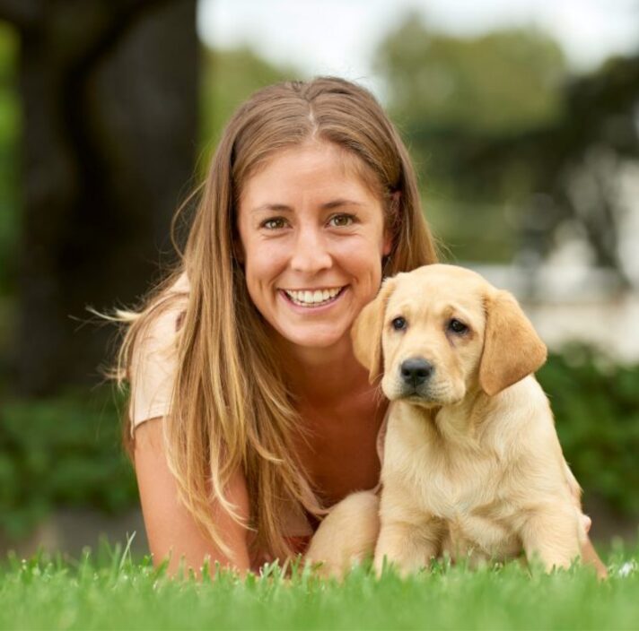 A person laying on some grass with an eight week old yellow labrador puppy. The person is smiling and looking at the camera. The puppy is also looking at the camera.