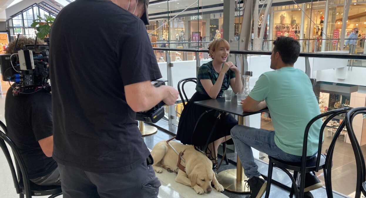 A cameraman filming Ingrid and Ryan at a cafe.