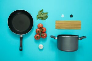 cooking equipment including a pot and pan. ingredients including tomatoes, onion & pasta. set on a light blue background