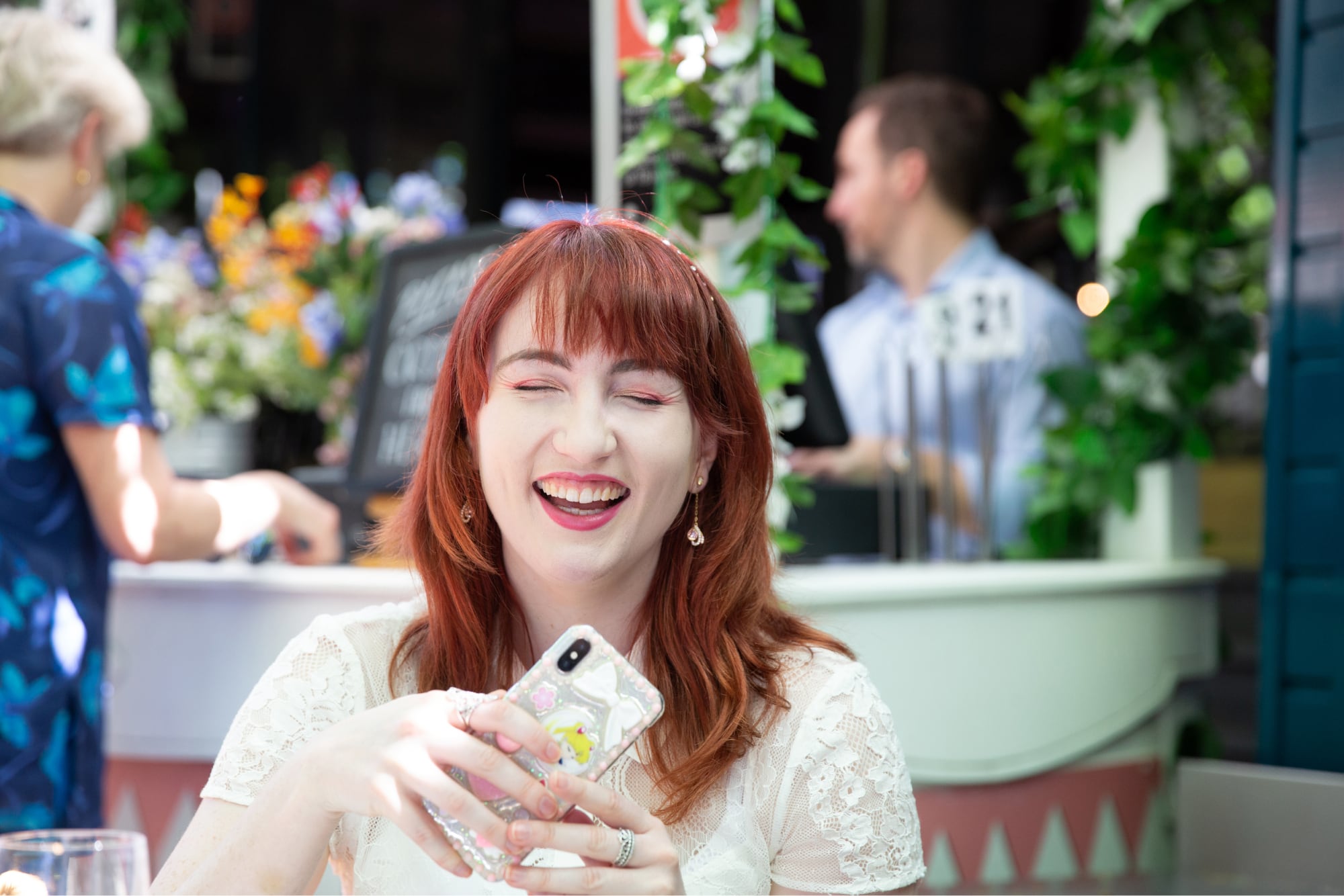 An adult sitting outside at a cafe laughing while holding their mobile phone.