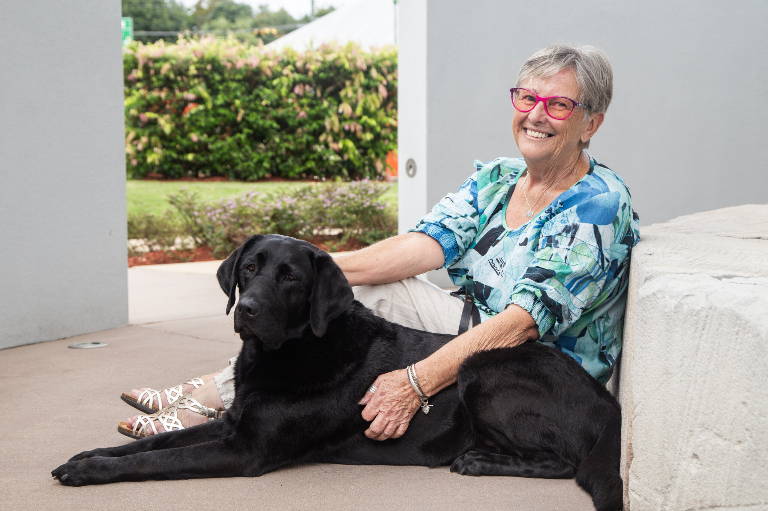 [Image description: Guide Dogs Client, Sue sits on the concrete outside with her hand on black Guide Dog, Vegas.]