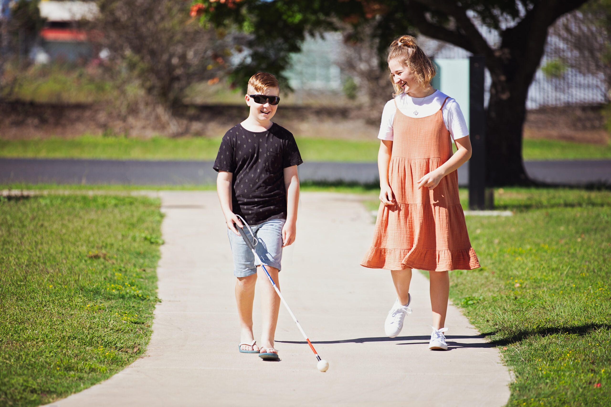 Two people walk down a path outside. One is using a White Cane.