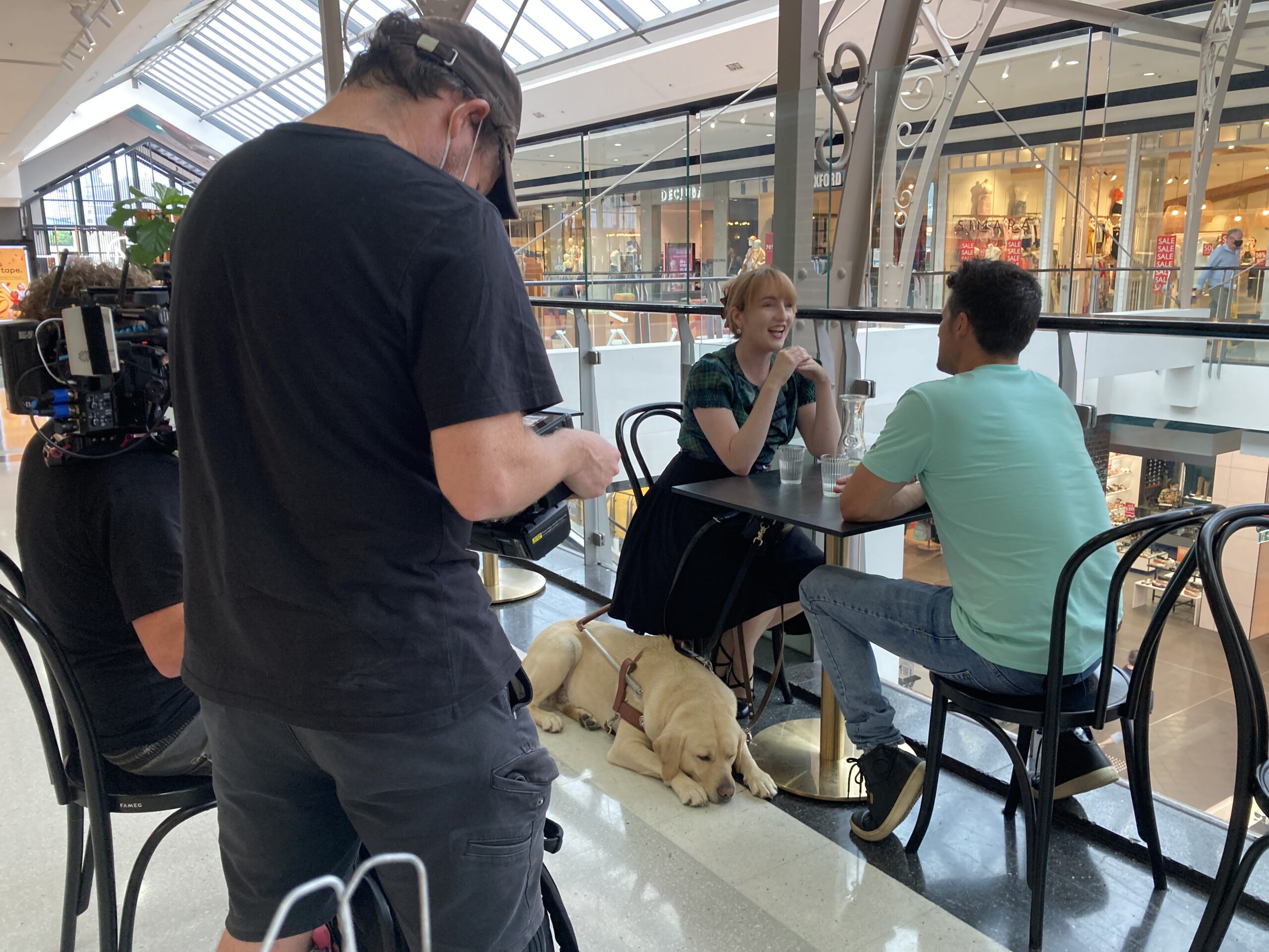 A cameraman filming Ingrid and Ryan at a cafe.
