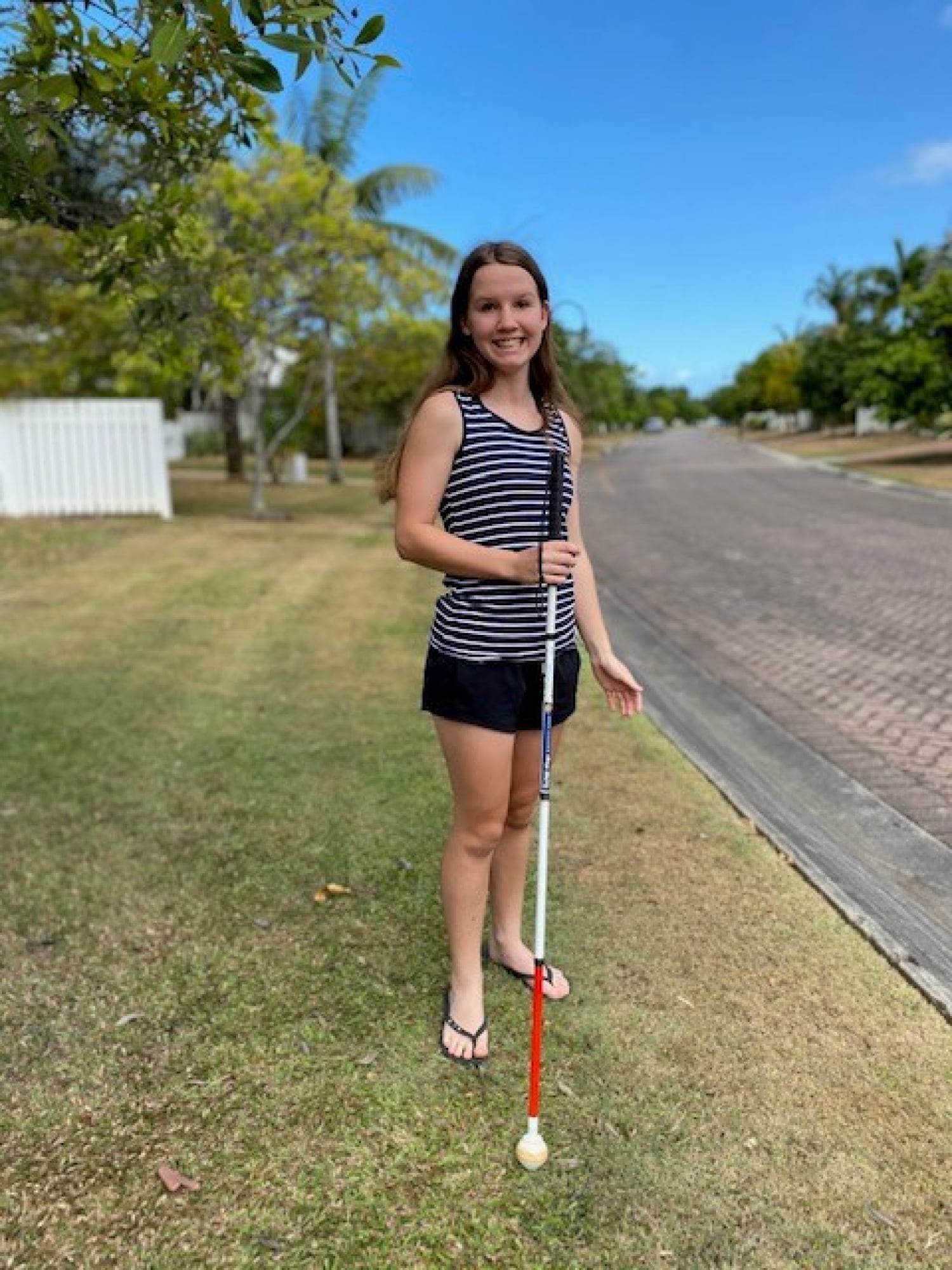 A young adult standing outside with their white cane. They are looking at the camera smiling.