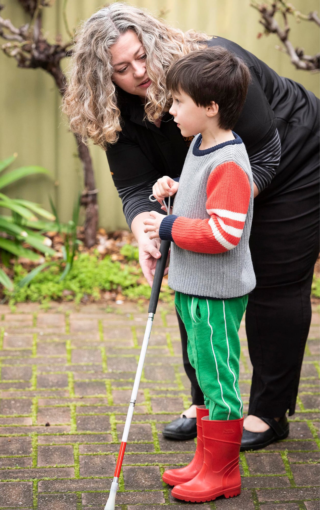 A young child using their white cane outside while an adult assists them.