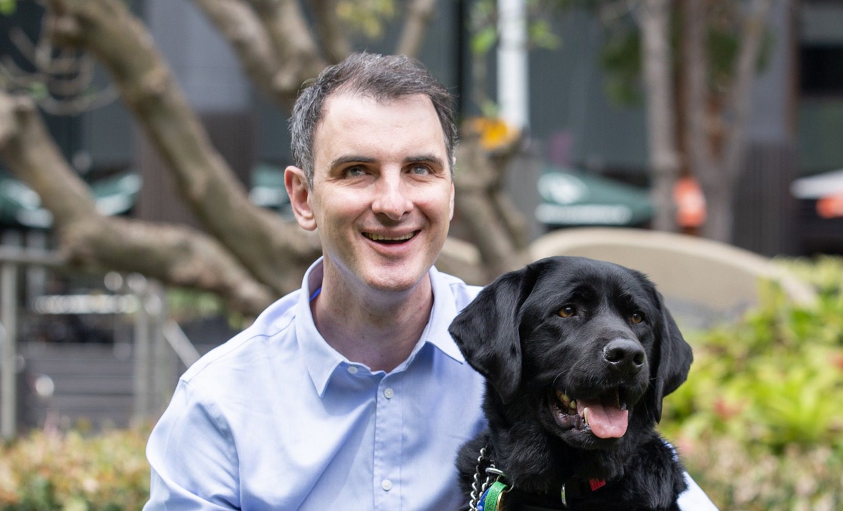 A man in a blue button up shirt and a black labrador are sitting in a grassy field. The labrador is wearing a Guide Dogs Queensland harness. Both are smiling at the camera.