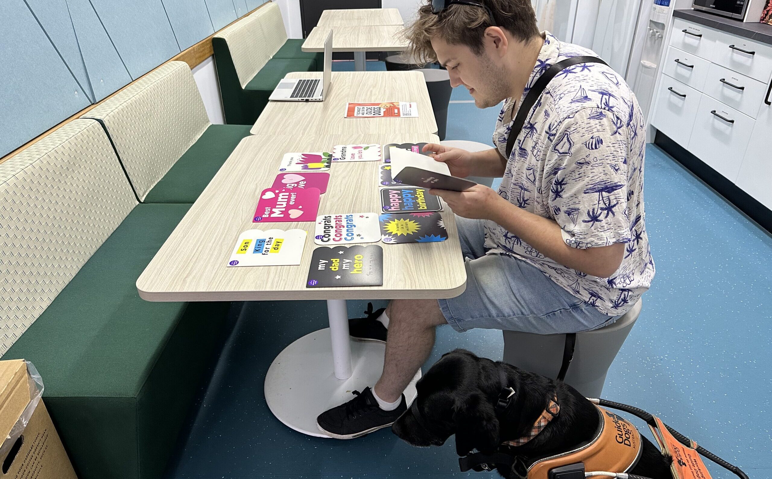 A male with low vision or blindness reads a braille card while sitting down at a table. Their Guide Dog sits at their feet.