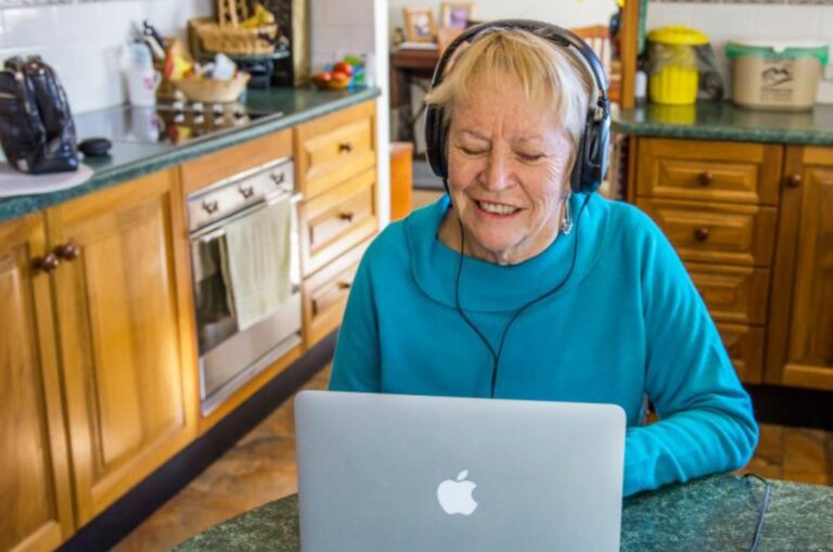 An older adult wearing headphones whilst on the laptop. The person is seated at a kitchen table.