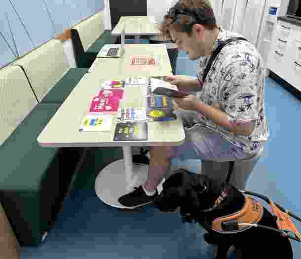 A male reads a braille birthday card sitting at a table. His Guide Dog sits beside him on the floor.