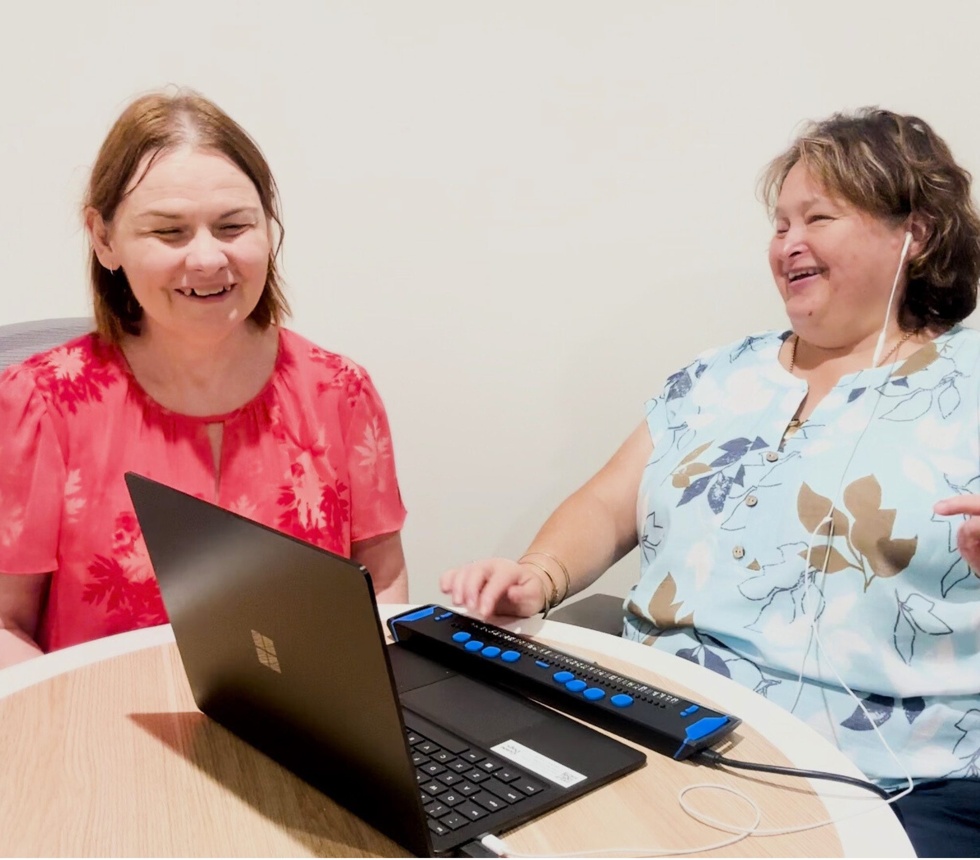 Two people laughing as they work together at a computer with a braille reader