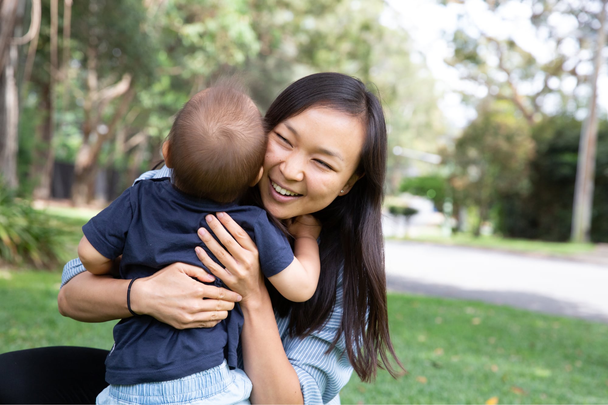A mother and baby outside on the grass hugging each other. The mother is smiling and the baby has its back to the camera.
