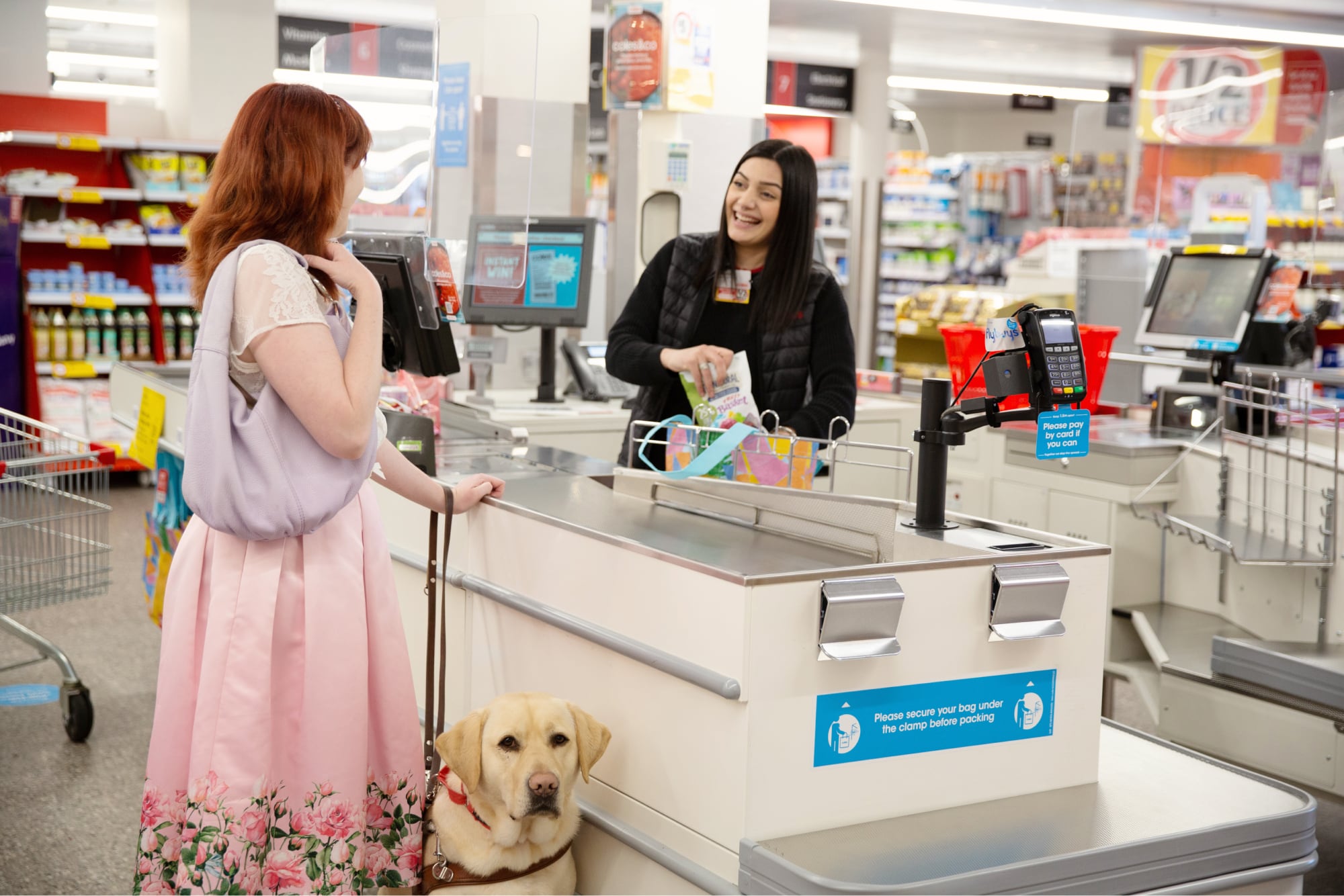 A person at a check out who is looking at the cashier. The cashier is smiling. The person at the checkout has a yellow labrador Guide Dog in harness who is looking at the camera.