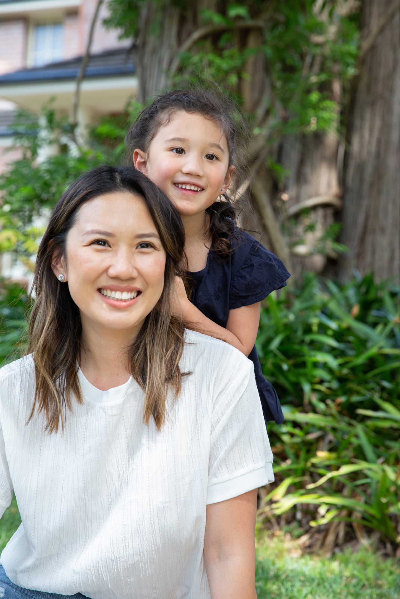 A mother and young child outside smiling at the camera. The young child is standing behind the mother with their hands on her back.