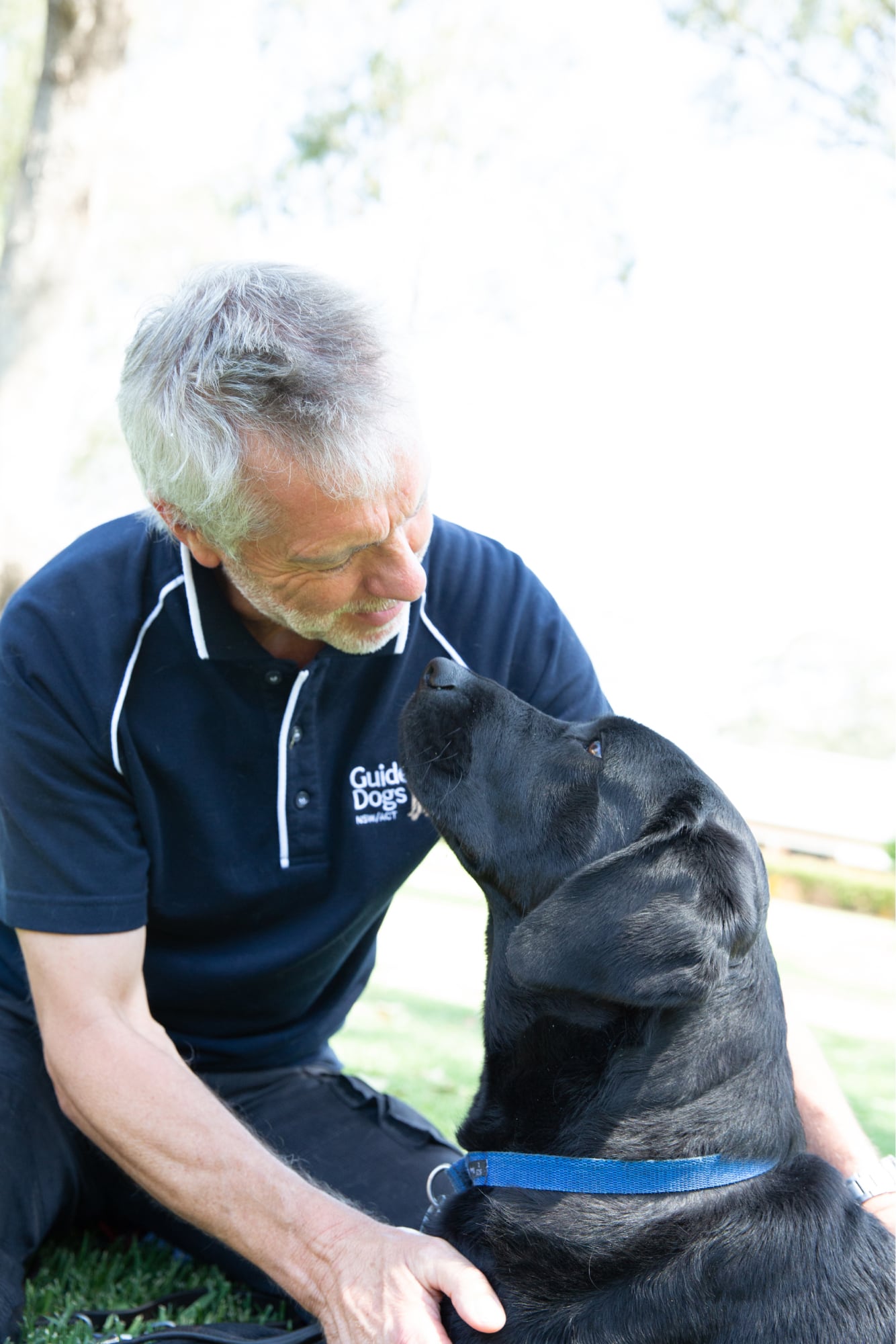 A Guide Dogs staff member and a black labrador dog sitting outside. The staff member and dog are both looking at each other.
