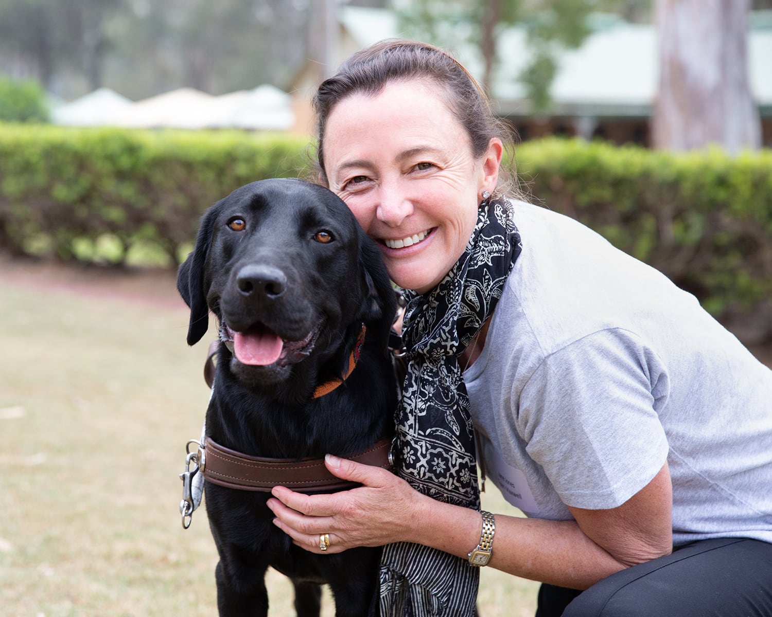 A person hugging a black labrador Guide Dog in harness. The person and dog are looking at the camera and the person is smiling,