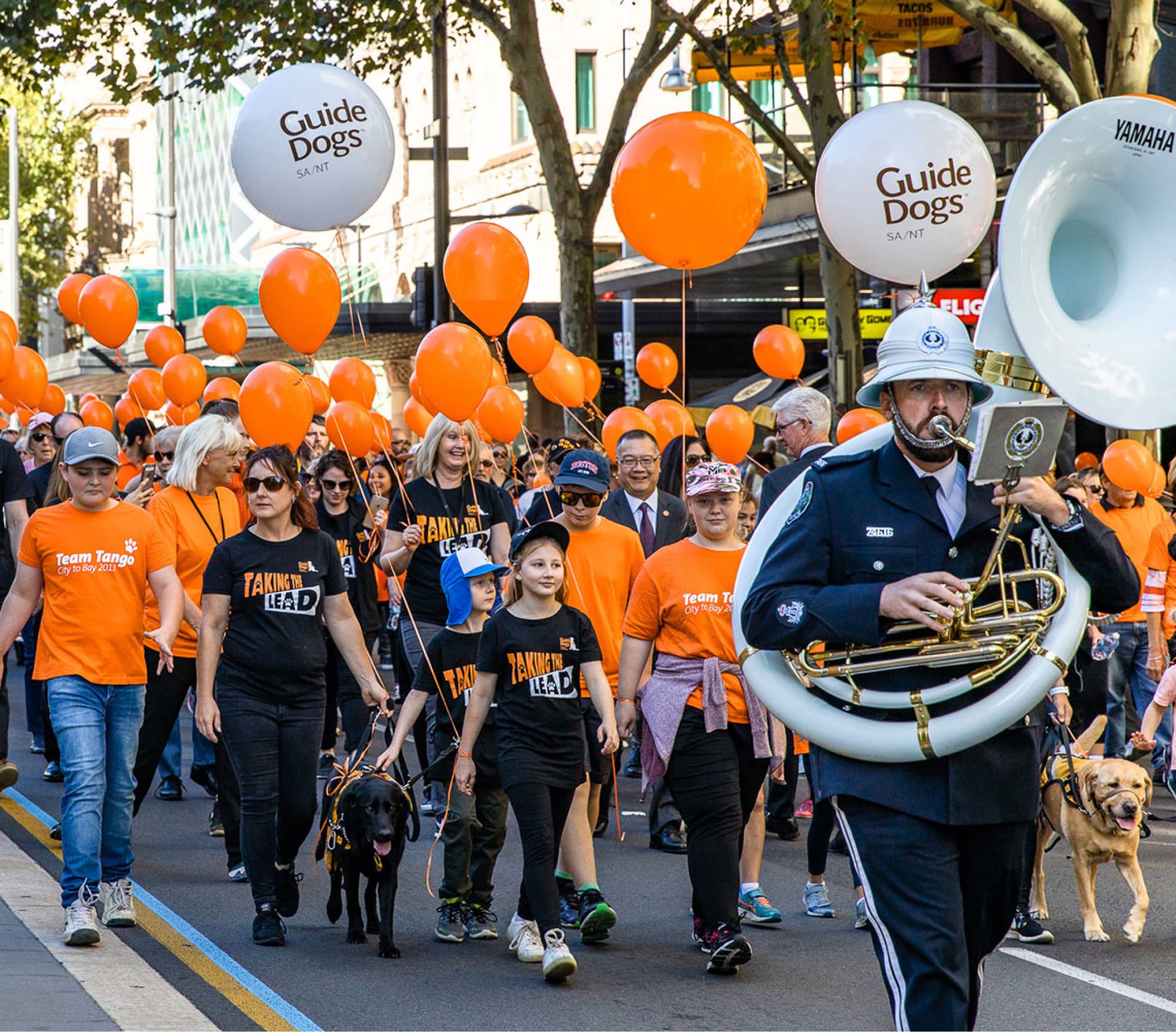 A large number of Guide Dogs staff and supporters walking in a parade. They are all wearing branded Guide Dogs orange t-shirts and some have orange branded helium balloons. At the front of the parade is a person playing a large french horn.
