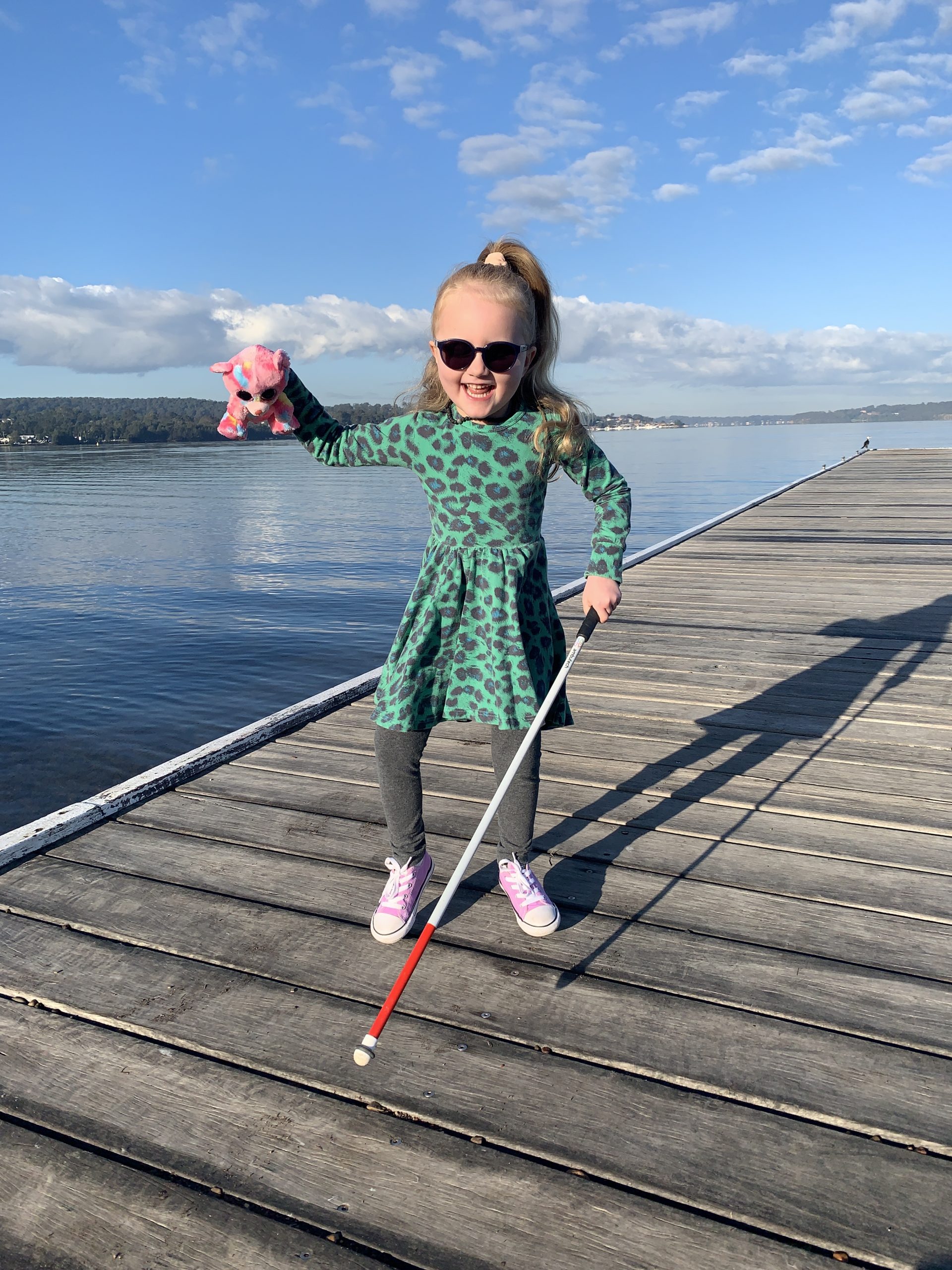 Stella holding her white cane in one hand and a toy in the other. She is standing on a boardwalk by the water.