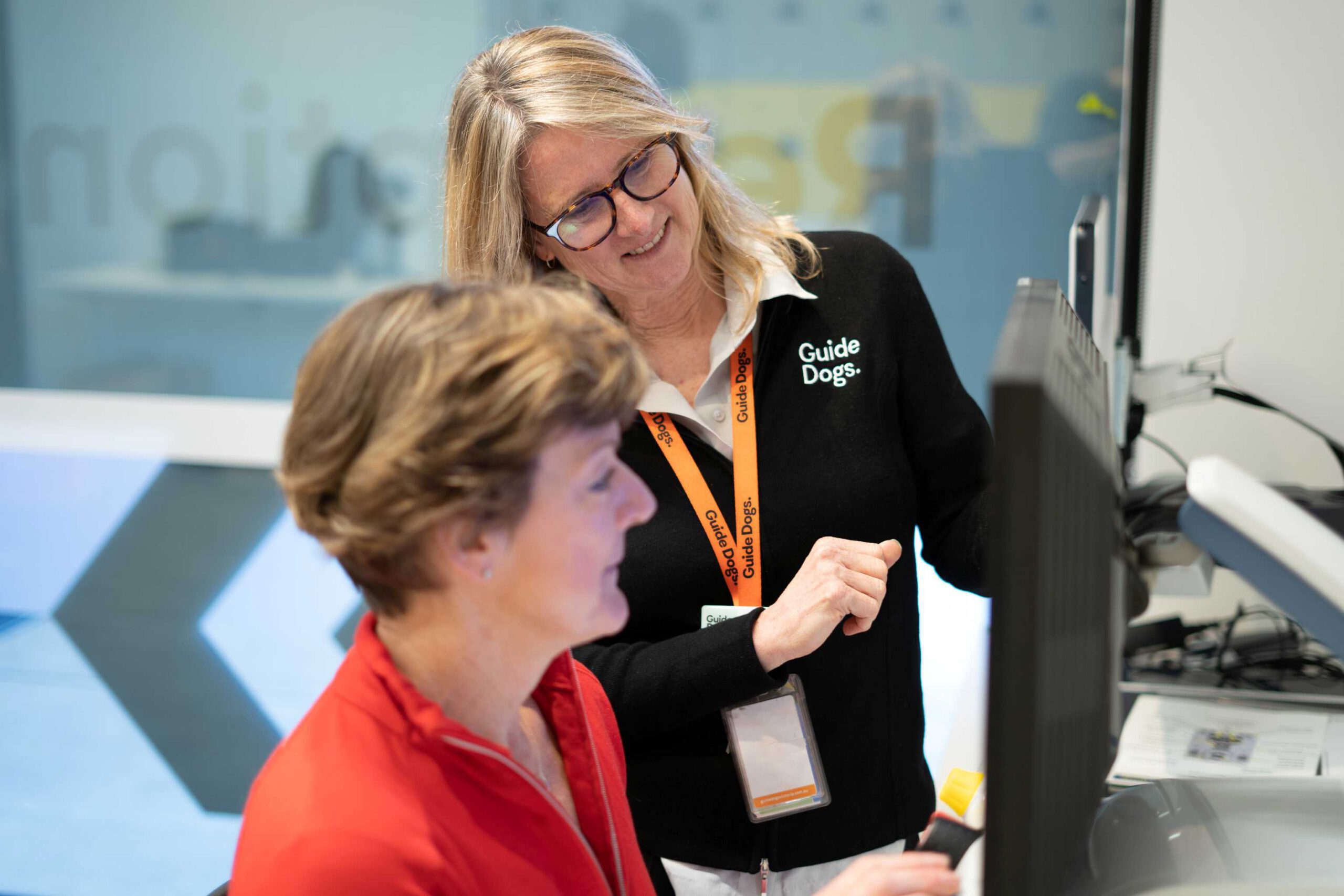 A person is sitting at a computer with a Guide Dogs staff member standing beside them. The Guide Dogs staff member is showing the person sitting at the computer how to it.