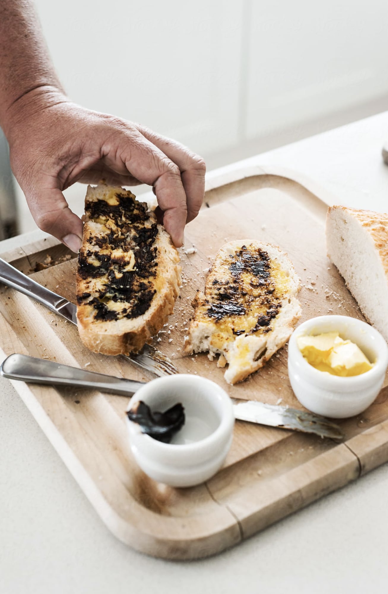 A person picking up a piece of buttered toast that is on a wooden board. There is a second piece of toast on the board