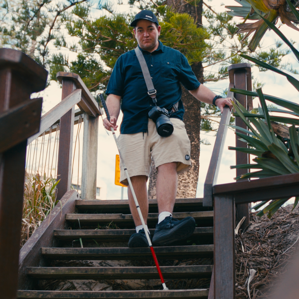 James wearing a cap and dark shirt, holds a white cane while descending wooden outdoor stairs, with a camera slung over his shoulder.