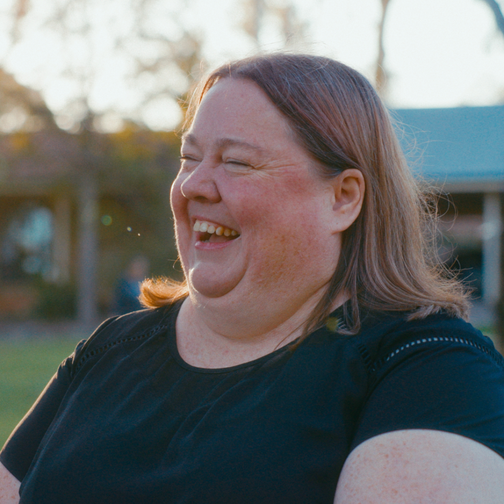 A side-profile portrait of Melissa laughing outdoors in the warm sunlight, wearing a black shirt.